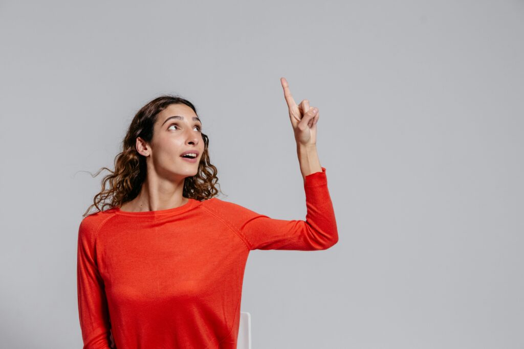 Portrait of a woman in red pointing upwards against a gray background, looking curious.